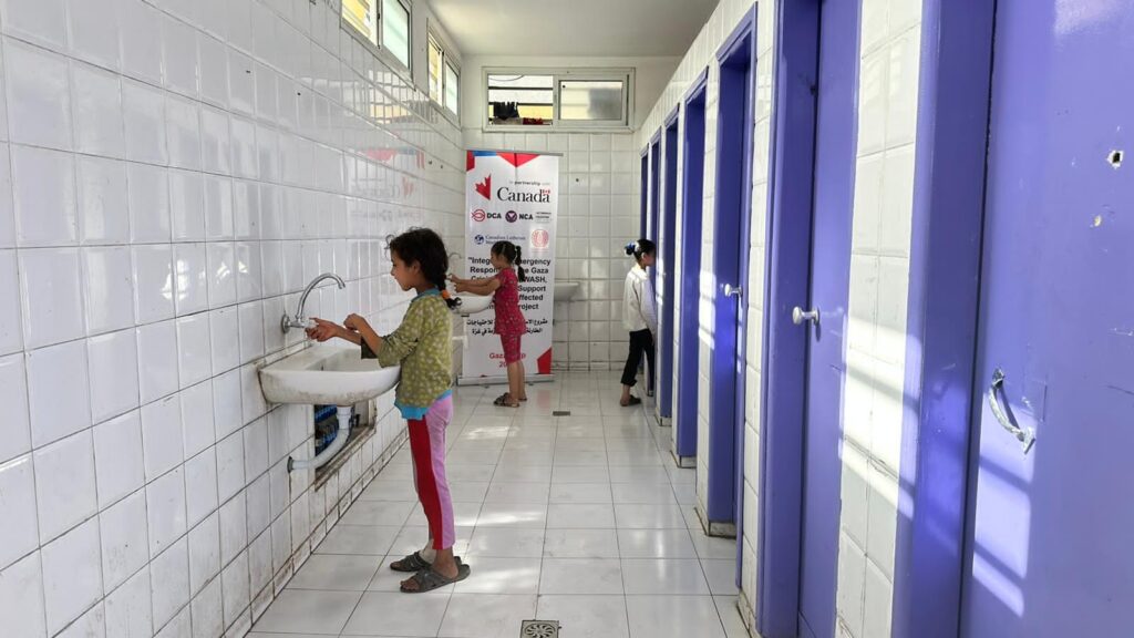 Two young girls washing hands in a public washroom, a third girl is entering a stall. 